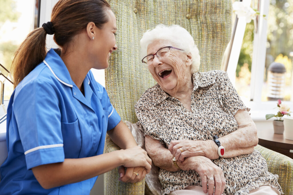 Senior Woman Sitting In Chair And Laughing With Nurse In Retirement Home Senior Woman Sitting In Chair And Laughing With Nurse In Retirement Home