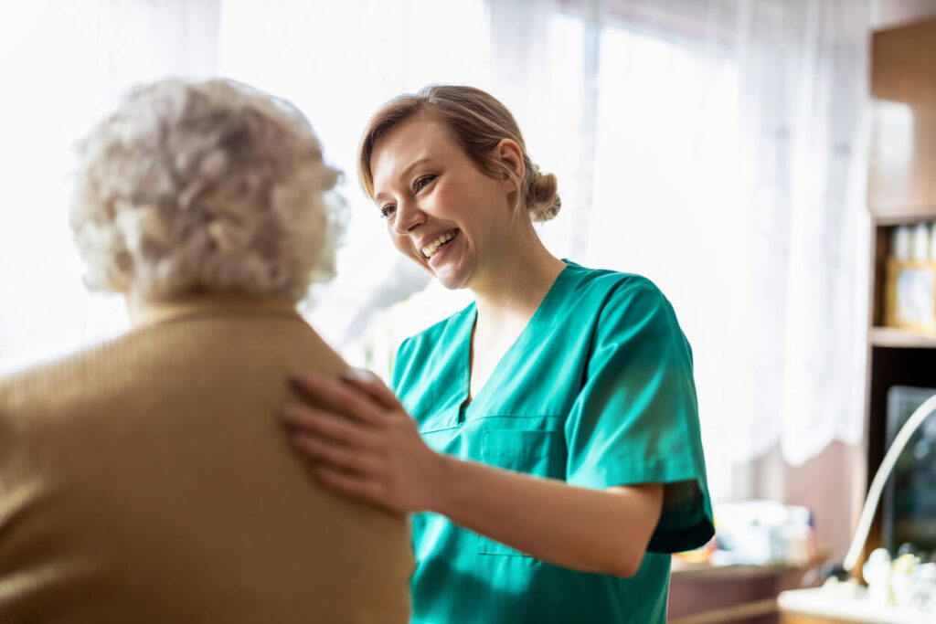 iStock-1307432645-2-1 A cheerful caregiver in green scrubs comforts an elderly person, placing a supportive hand on their shoulder in a bright room|A caregiver in blue scrubs assists an elderly person standing up from a couch